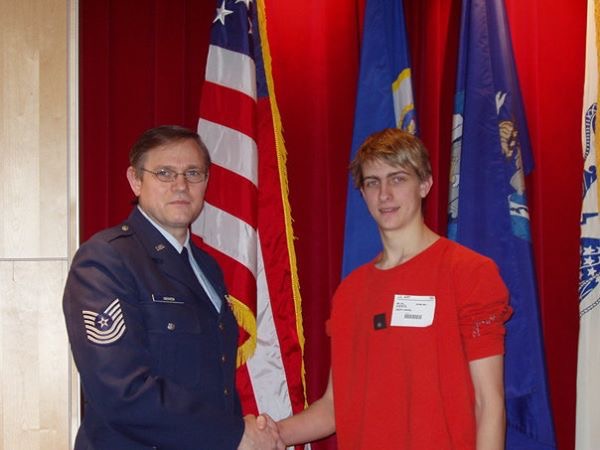 Stephen Groven dressed in his military dress blues shaking his son's hand with flags in the background