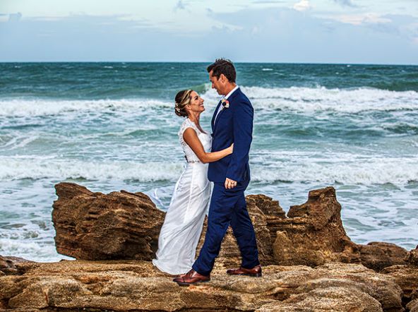 Andrew and Natasha Hidalgo celebrating their marriage on the rocks at the beach in their wedding attire