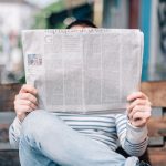 woman sitting on a park bench with her legs crossed reading a newspaper