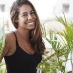 Carefree lovely tanned young woman standing balcony near green plant smiling laughing out loud look adorable grin camera talking casually coworker veranda, enjoy perfect sunny summer days.